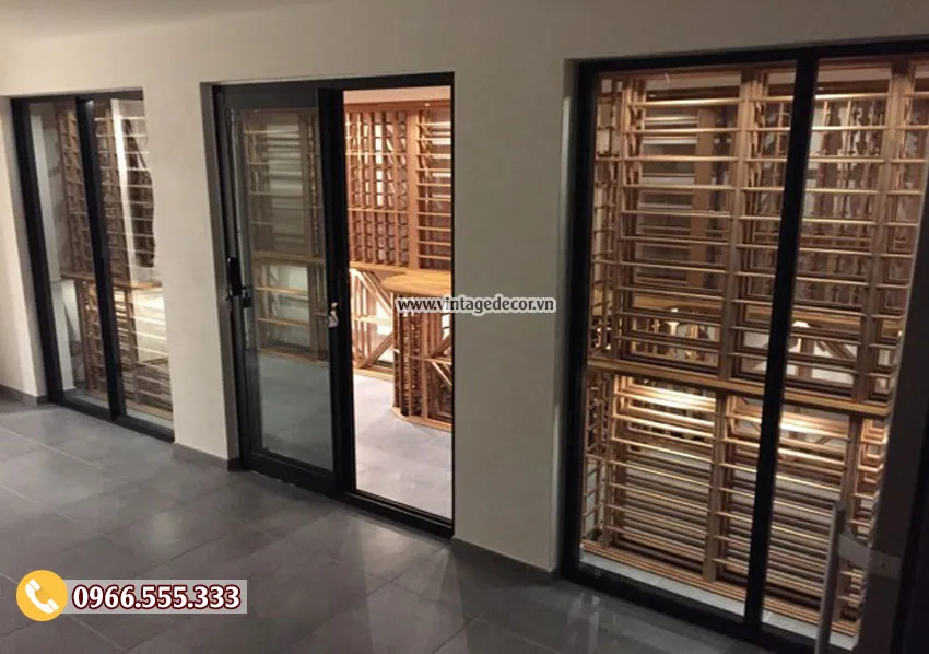 Cửa kính khung đen của hầm rượu mini HR94 nhìn từ hành lang, thấy kệ gỗ bên trong