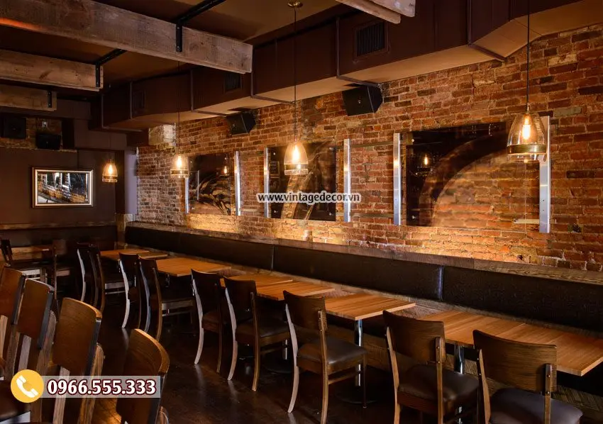 Dãy bàn gỗ dài tựa tường gạch thô trong quán bar BR47, đèn thả chụp kính