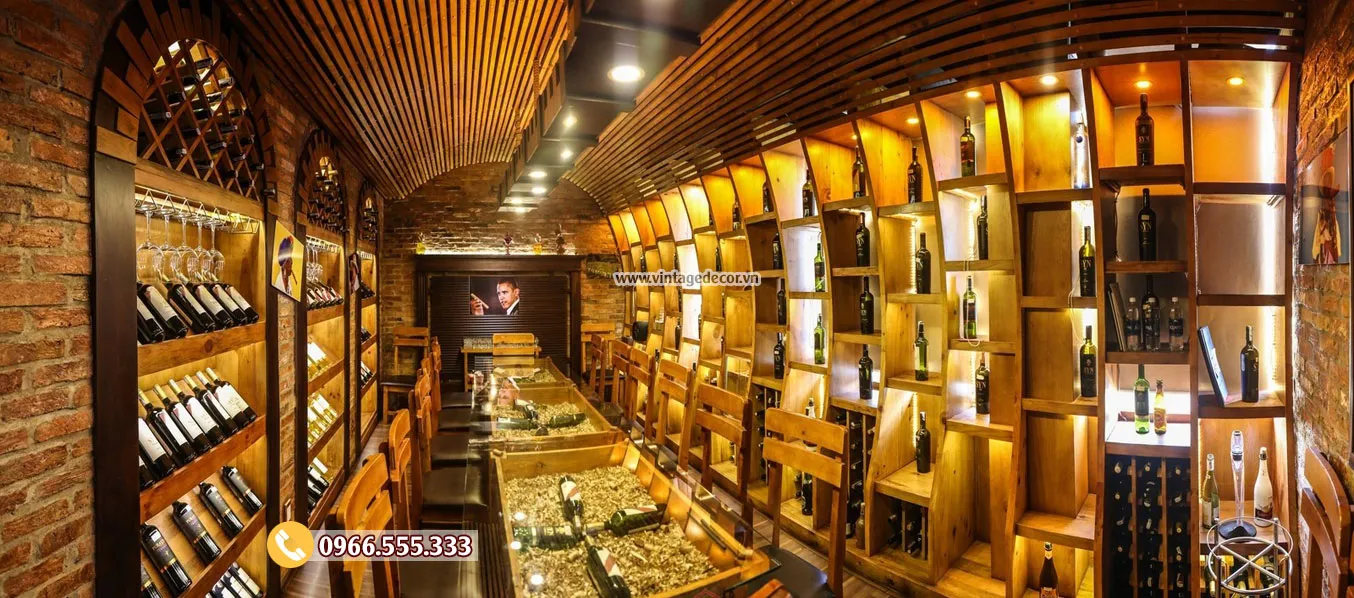 Phòng dài hẹp, một bên tường gạch có kệ ô chéo, một bên kệ ô vuông sáng đèn vàng, trần gỗ vòm