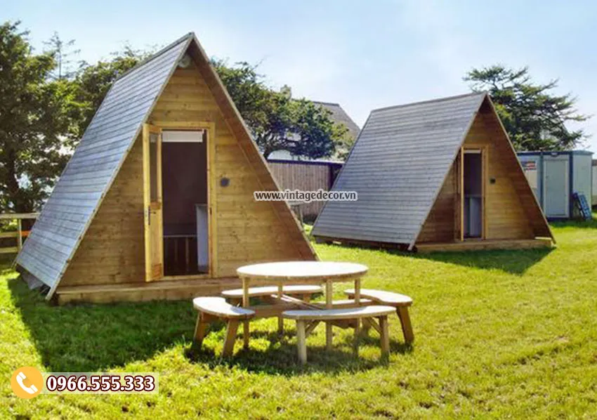 Hai căn bungalow mái chữ A lợp tấm xám đặt trên bãi cỏ, phía trước có bàn tròn và ghế băng gỗ