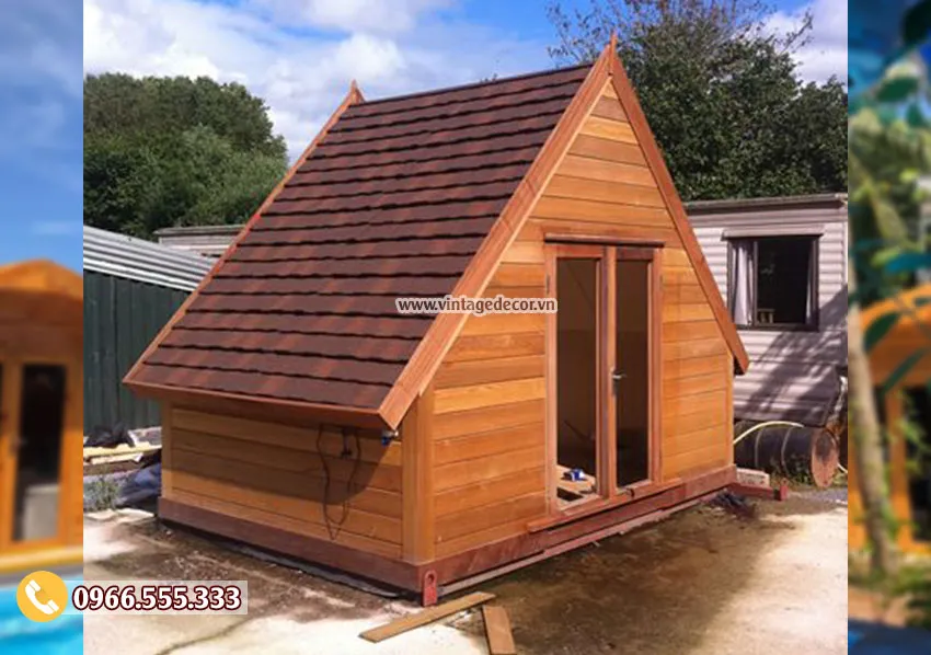 Bungalow mái tam giác lợp ngói bitum nâu, vách ván gỗ ngang, cửa kính đôi, đặt trên khung đế
