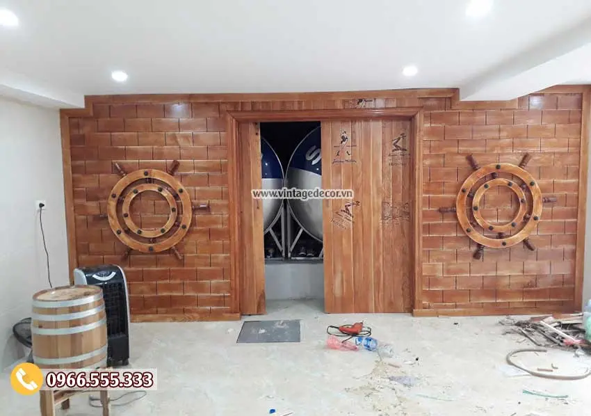 Bức vách ốp gỗ giả gạch có bánh lái tàu, cửa lùa mở ra thấy bồn nước phía sau