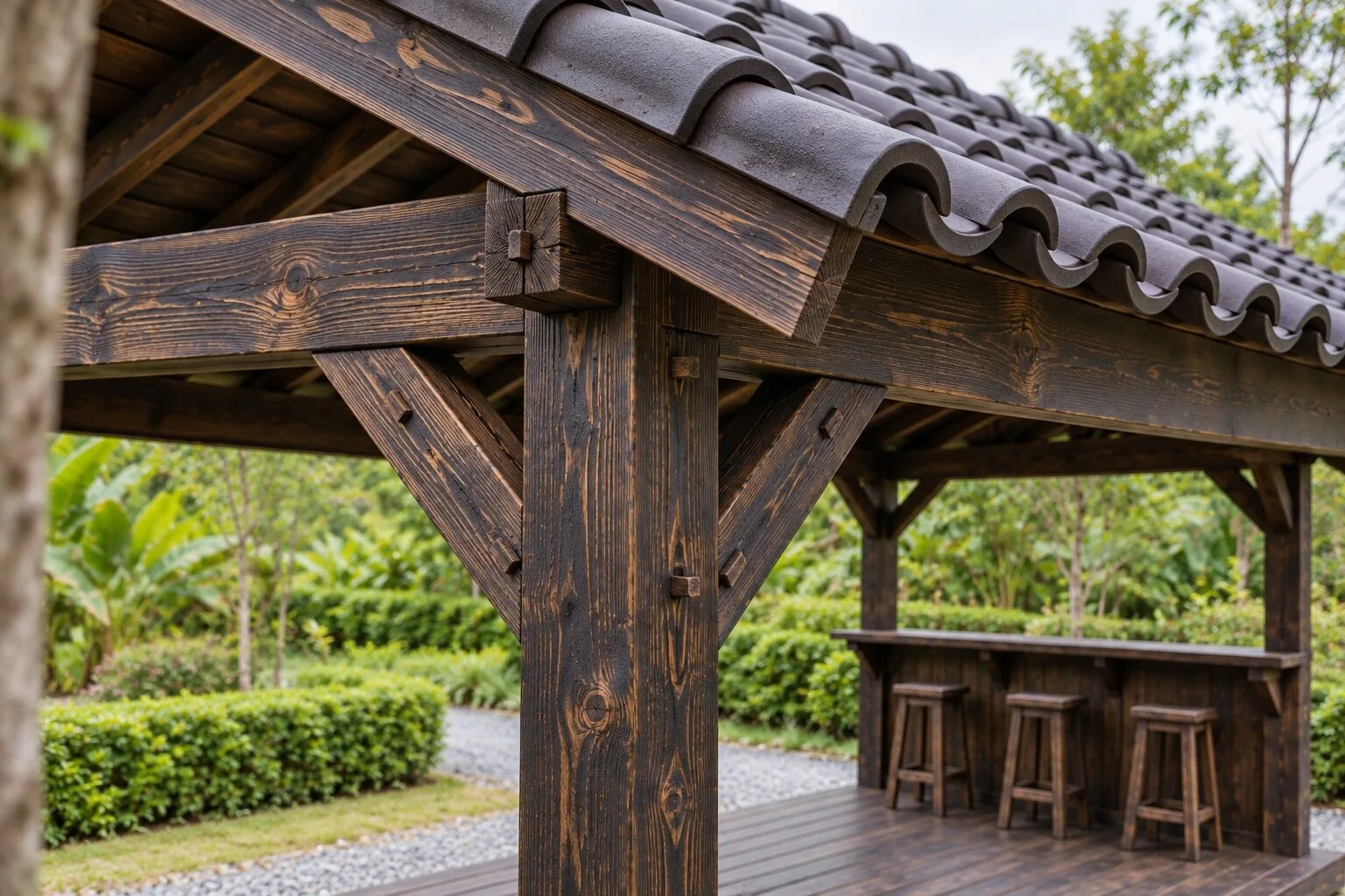Cận cảnh góc chòi T103: cột gỗ sẫm có thanh chống chéo đỡ dầm, mép mái ngói đua ra, phía sau là quầy ván đứng và ba ghế đẩu trên sàn gỗ